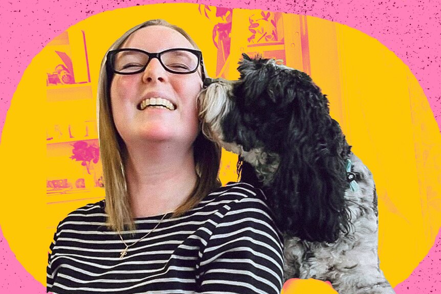A young woman wearing glasses and a stripey top is being kissed on the cheek by her cute black dog