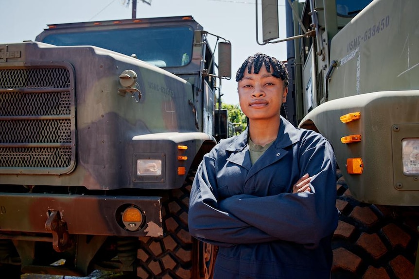 A woman with her arms crossed in front of her, standing between two large army trucks