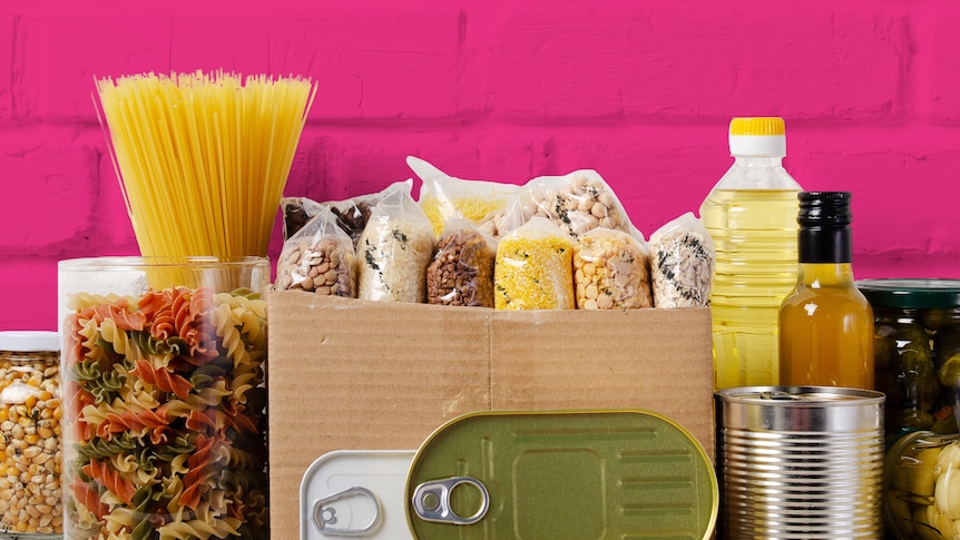 Various pastas, grains, tinned foods and bottled oils are seen in front of a pink brick background.