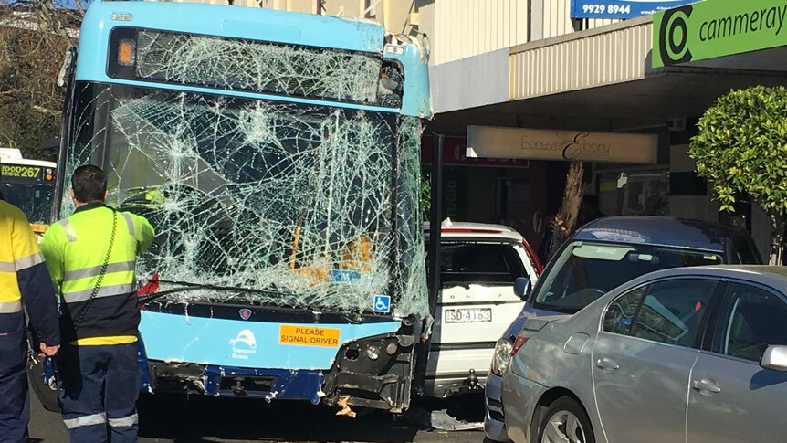 A bus with a smashed screen and broken bumper on the street and a car across the footpath, with emergency workers looking on