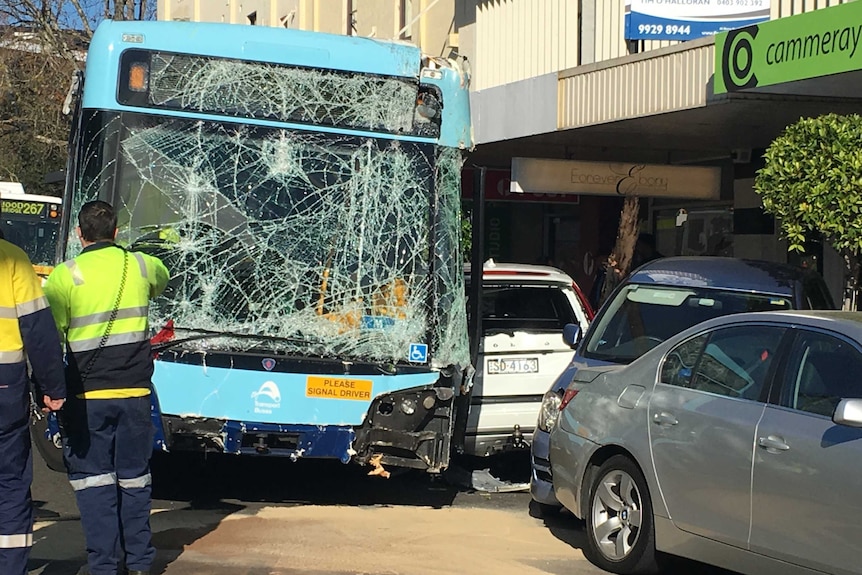 A bus with a smashed screen and broken bumper on the street and a car across the footpath, with emergency workers looking on