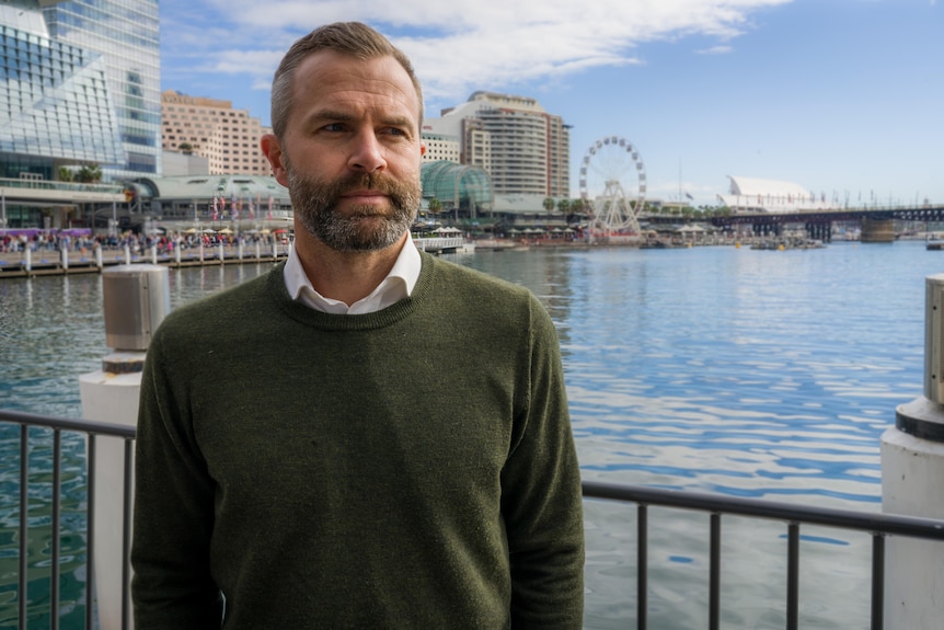 Man with trimmed beard and cropped hair wearing a green pullover and white shirt, with Darling Harbour behind