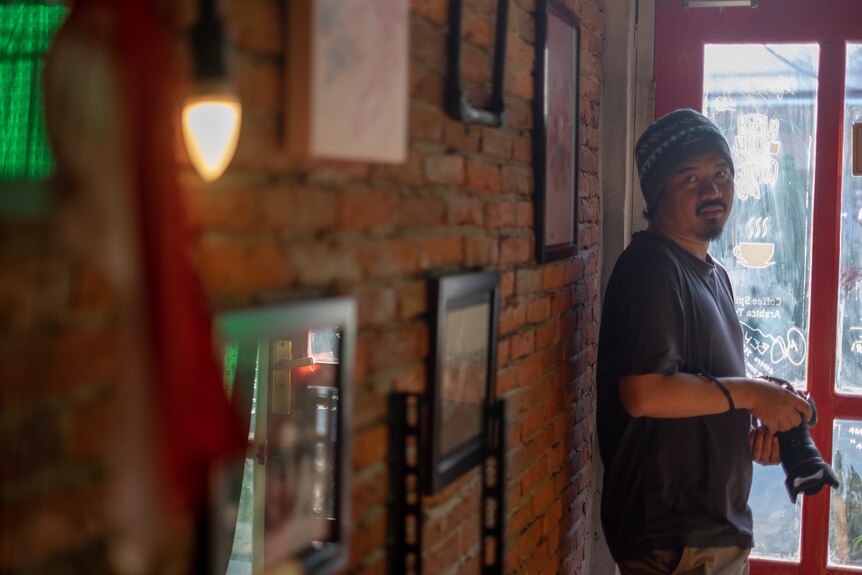 A Indonesian man wearing a warm hat stands near a doorway holding a camera.