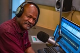 A picture of a man wearing a maroon shirt and headphones with a radio microphone and a computer screen showing audio wavelengths