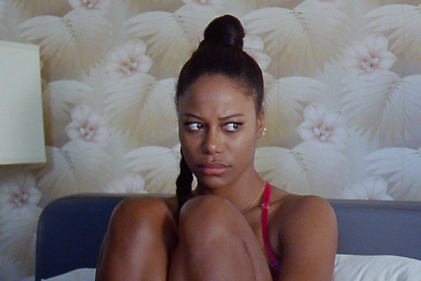 Young black woman with dark hair in high ponytail wears a red crop top and sits on a hotel room bed looking surly.