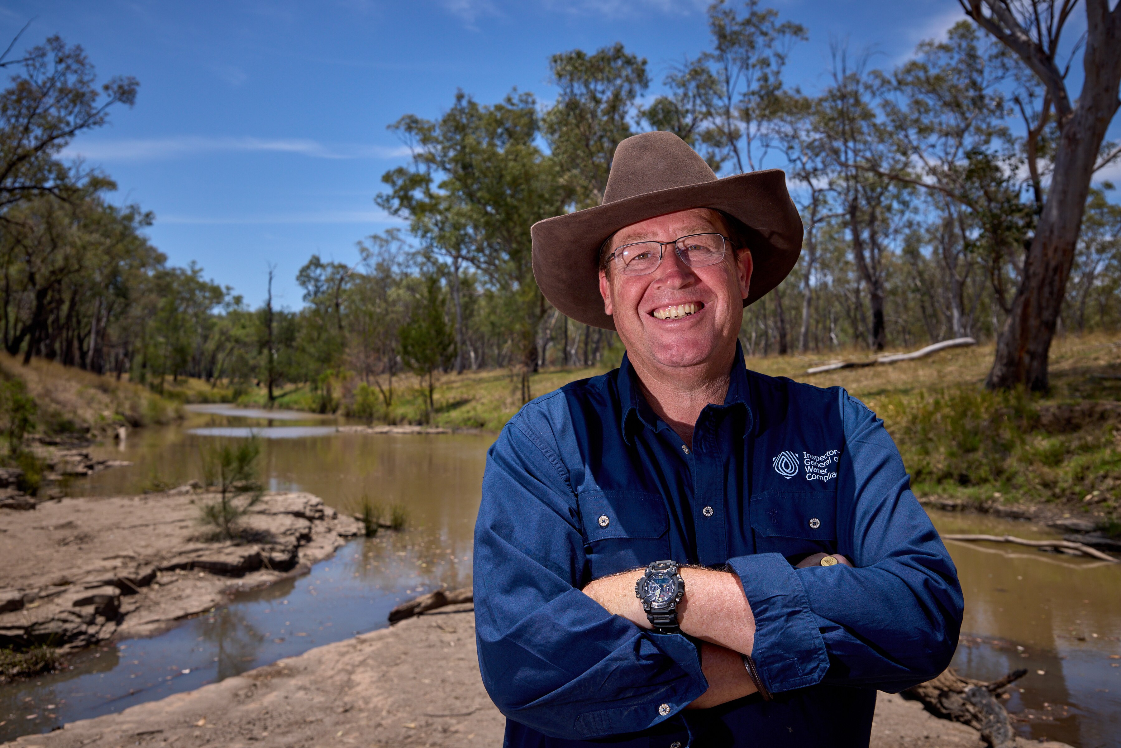 Troy Grant viste un cuello abotonado de manga larga azul oscuro y un sombrero marrón. Está parado frente a un río y árboles de caucho.