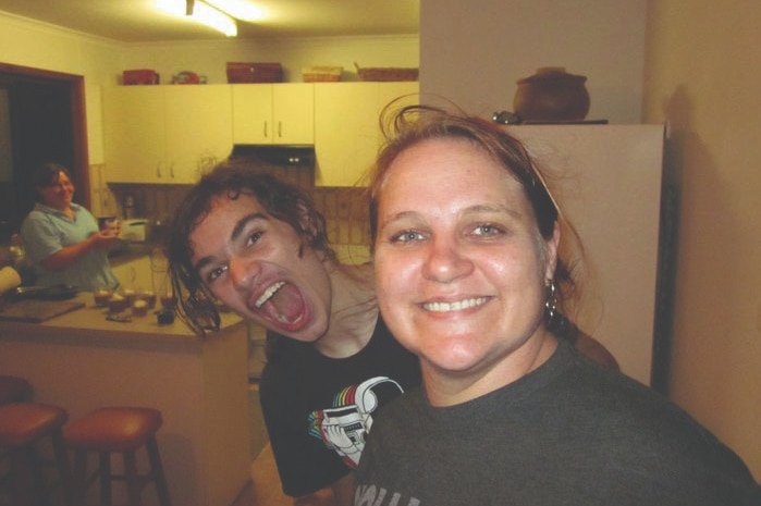 A woman with blue eyes and brown hair smiles at the camera, a young teen boy pulls a funny face at the camera behind her