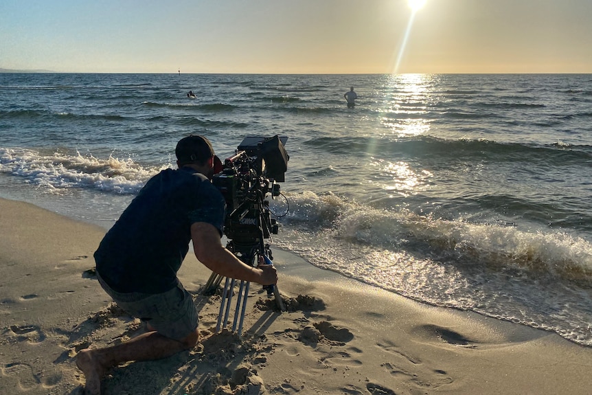 Camera operator on a beach filming a woman standing in water out at sea.