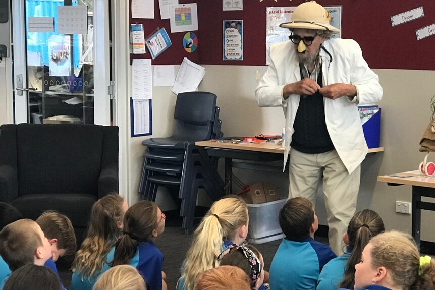 A man in a hat, glasses and a fake nose stands in front of a class of primary school children.