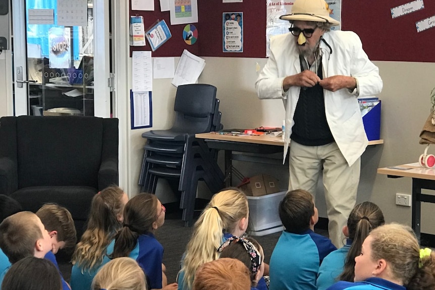 A man in a hat, glasses and a fake nose stands in front of a class of primary school children.