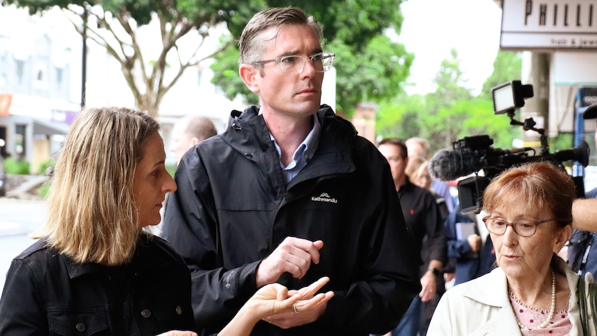 Two women and a man walking together outdoors, with a crowd of people and cameras in the background.