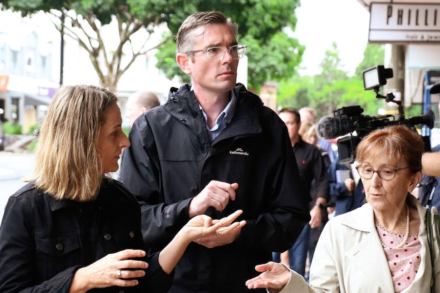 Two women and a man walking together outdoors, with a crowd of people and cameras in the background.