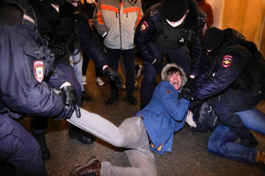 Russian police officers hold the limbs of a protester who is lying on the ground with a distressed expression.
