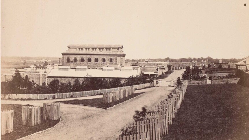 A black and white photograph of the outside of a brick building with perimeter wall.