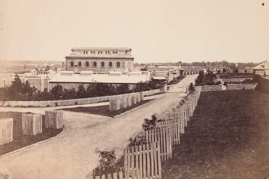 A black and white photograph of the outside of a brick building with perimeter wall.