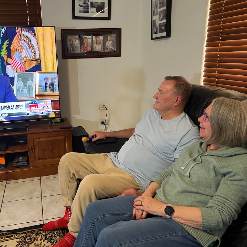 A middle aged couple watch Fox News coverage of the aftermath of the Trump shooting in their living room.