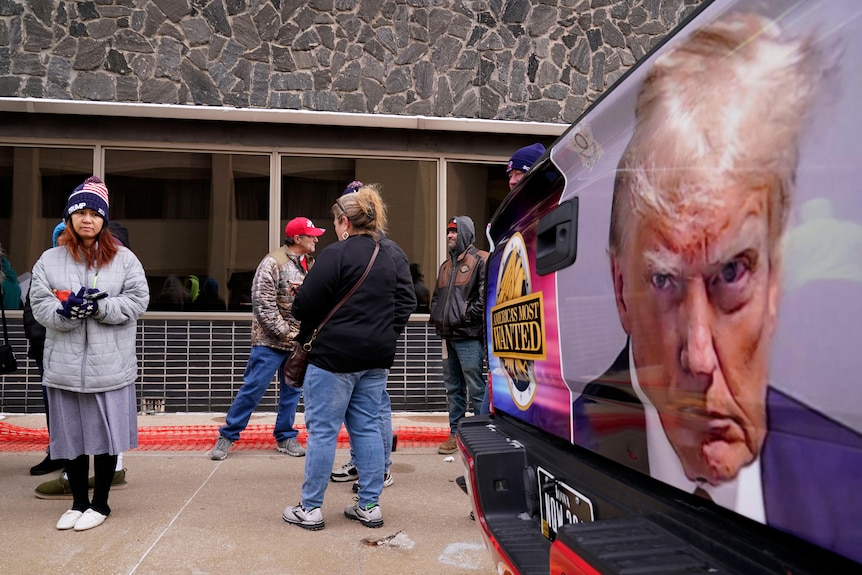 A group of people wearing cold-weather clothing and Donald Trump-branded hats stand beside a large poster of Mr Trump's mugshot.