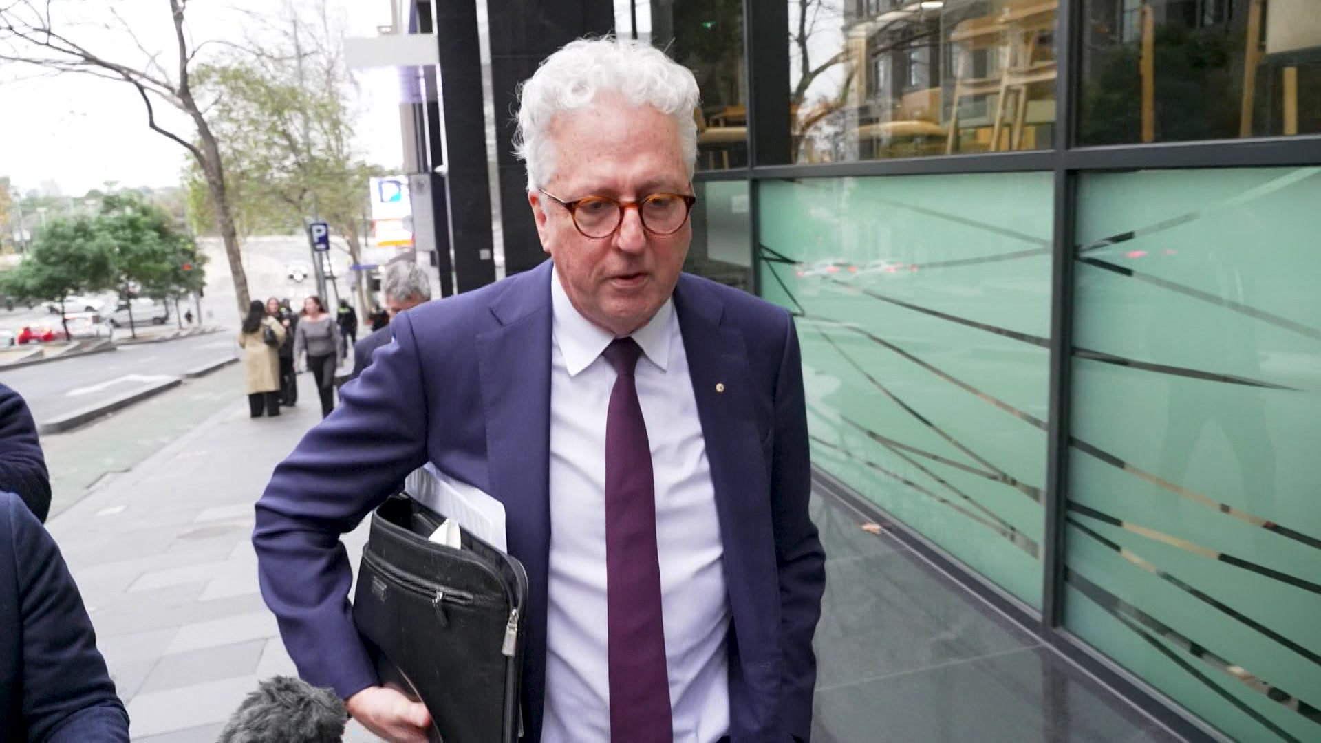 A white haired man with glasses in a blue suit and red tie walks along a footpath with a briefcase under one arm.