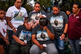 A group of people, some wearing shirts featuring the photo of a young girl, hold candles and weep.