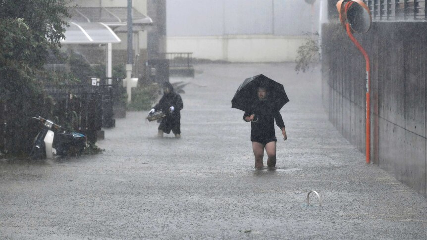 Two mean with their trousers rolled up wade through a flooded street, carrying household items and an umnbrella.