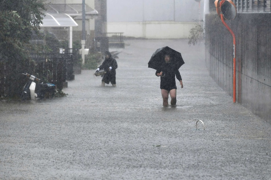 Two mean with their trousers rolled up wade through a flooded street, carrying household items and an umnbrella.