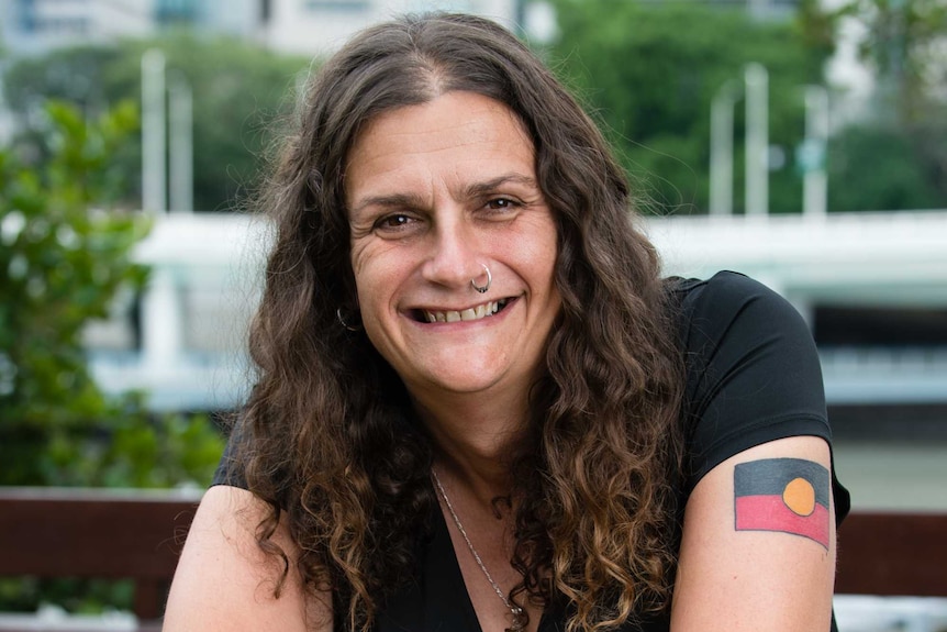 Woman with long dark wavy hair, and a Aboriginal flag tattoo, smiles at the camera.