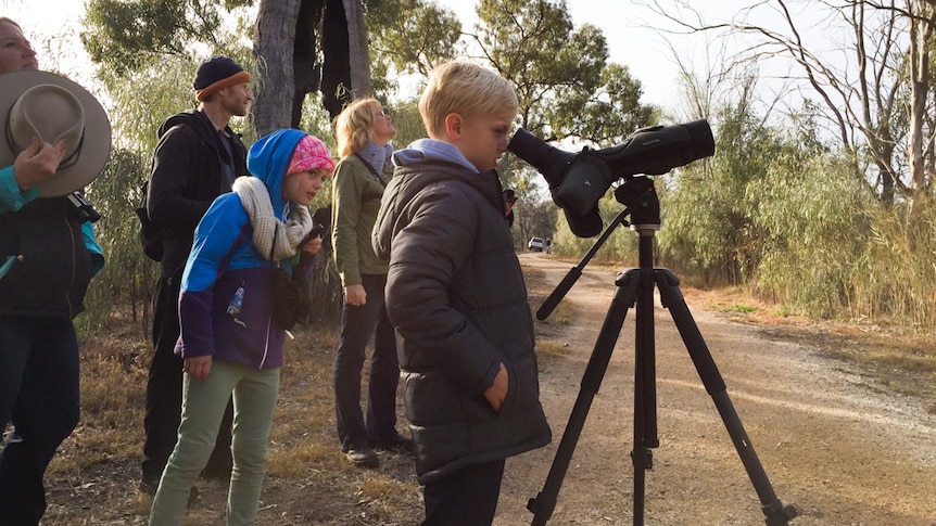 Declan May looks through a telescope surrounded by people in a native vegetation setting.