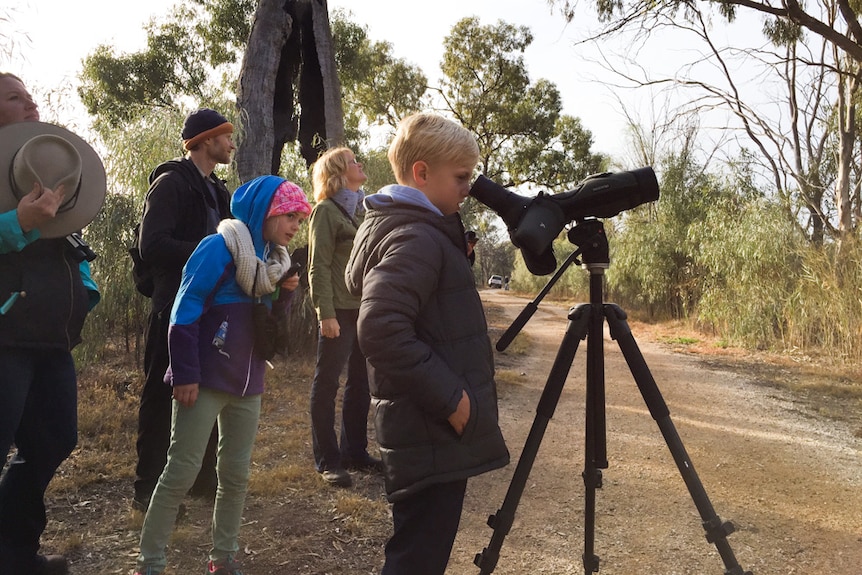 Declan May looks through a telescope surrounded by people in a native vegetation setting.