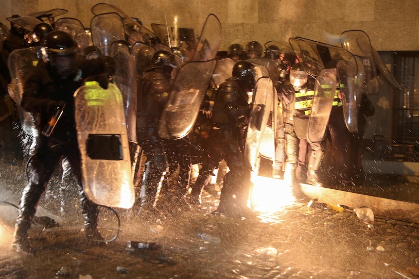 A group of police in riot gear react to a flame on the ground caused by a molotov cocktail