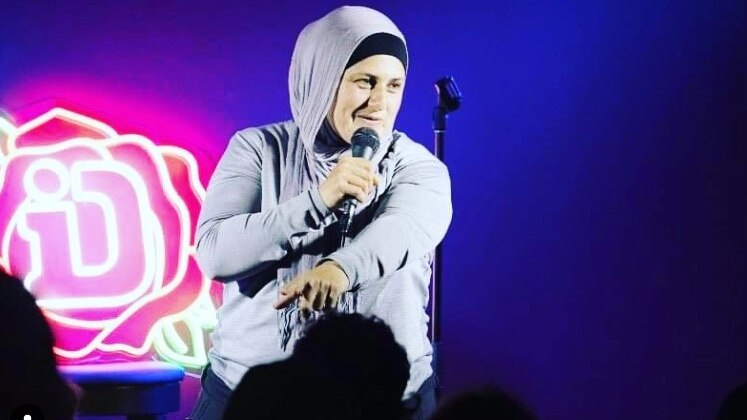 Frida Deguise smiling on stage holding microphone, wearing hijab, with bright purple light behind her.
