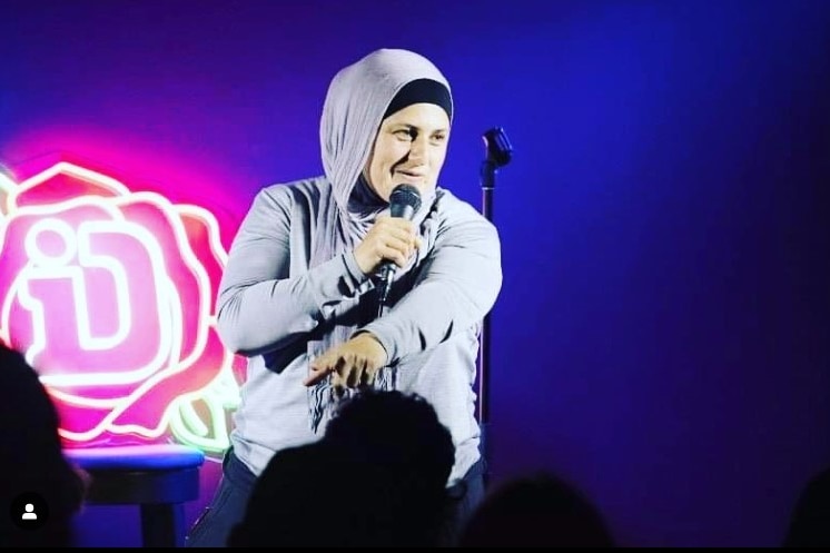 Frida Deguise smiling on stage holding microphone, wearing hijab, with bright purple light behind her.