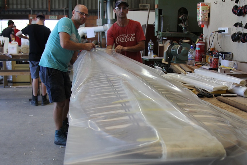 Two men push a surfboard into a clear plastic bag.
