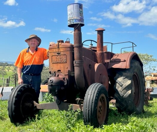 A middle-aged man in a hi-vis shirt stands next to a rusty vintage tractor on a rural property.
