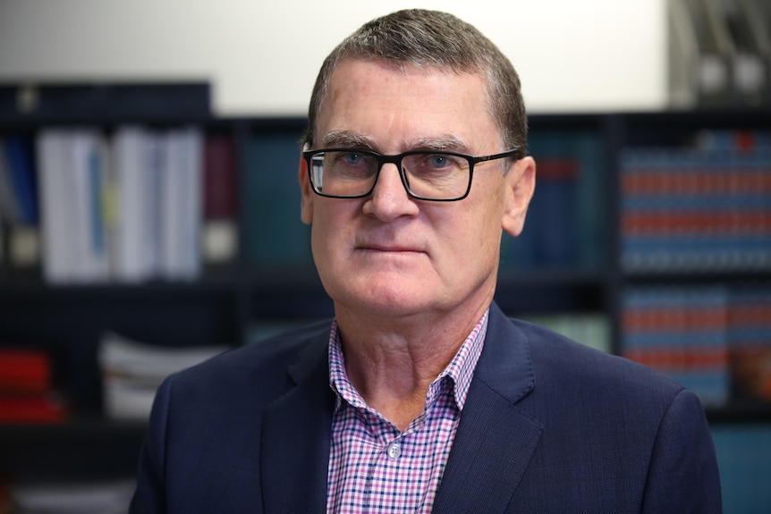 A man in a jacket and shirt with glasses looks serious. 