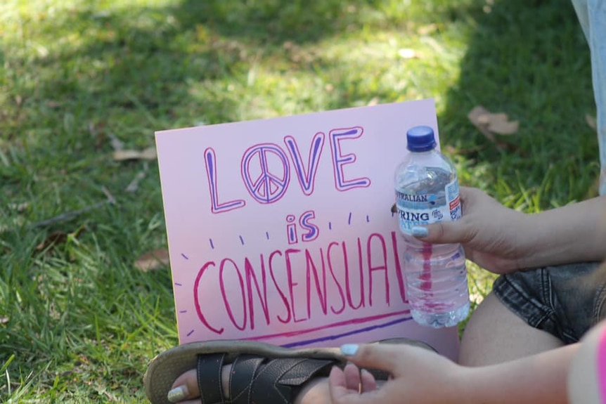 A small pink cardboard sign reading 'Love is Consensual', held by someone out of frame sitting on grass
