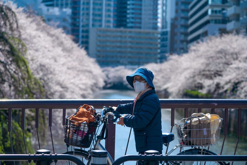 An older Japanese woman in a face mask wheeling her bike over a bridge