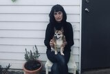A woman with black hair waring a black jumper sitting on her front porch with a dog on her lap.