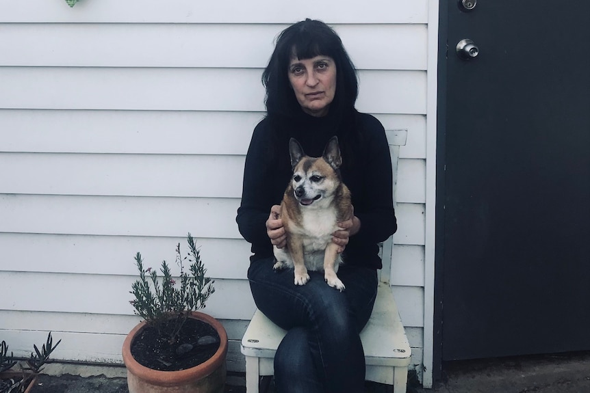 A woman with black hair waring a black jumper sitting on her front porch with a dog on her lap.