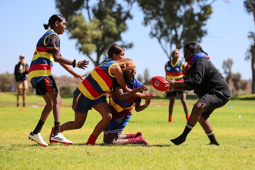 A group of young Aboriginal women wearing football jumpers compete for a ball on the ground in the middle of the day
