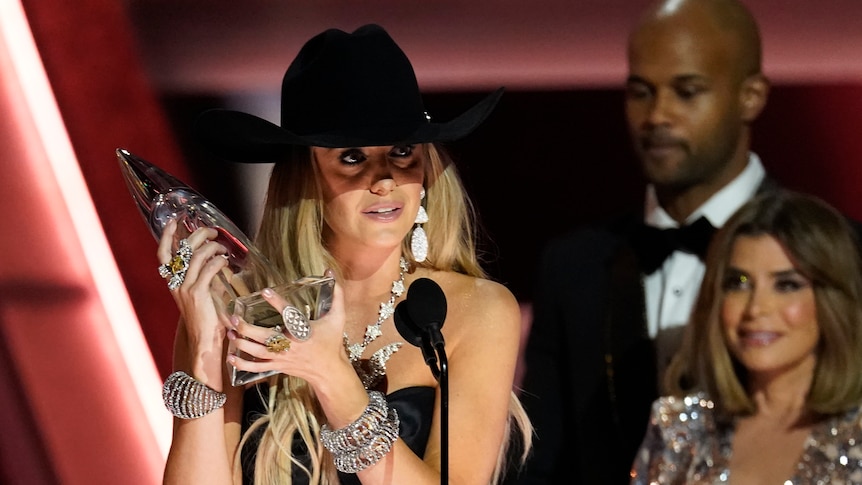 A young white woman with long blonde hair in a black cowboy hat holds a clear glass trophy as she speaks behind a lectern.