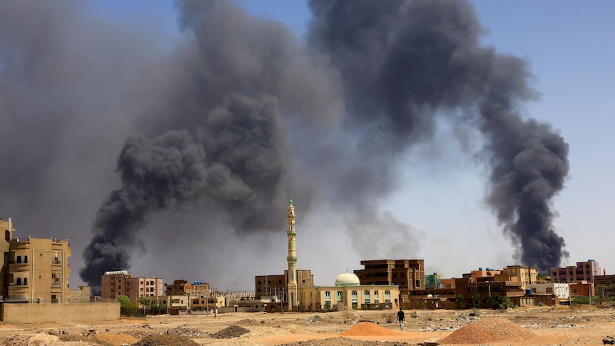Smoke rises from buildings in the city of Khartoum, where a man can be seen in the foreground walking on the ground.
