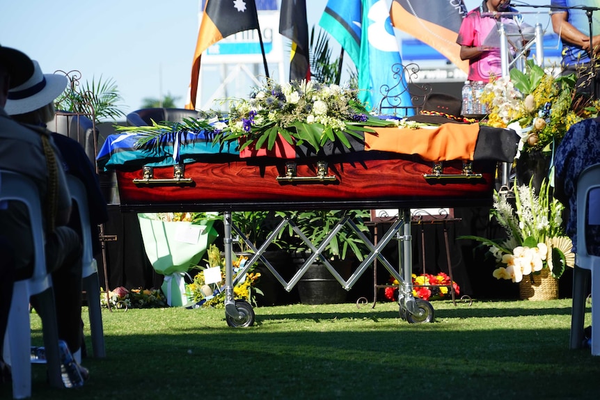 A coffin covered with flags and flowers can be seen. It is made of wood.