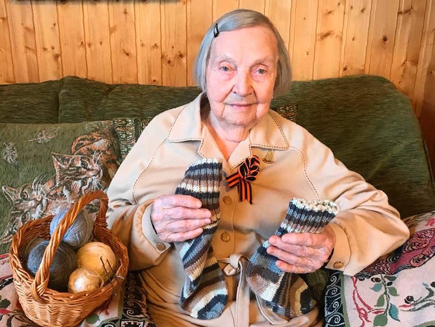 An old woman sits on a green couch holding a pair of knitted socks. A basket of yarn sits beside her