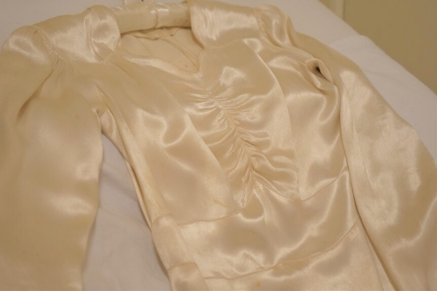 The bodice of a cream satin long-sleeve wedding dress lies on a white surface. 
