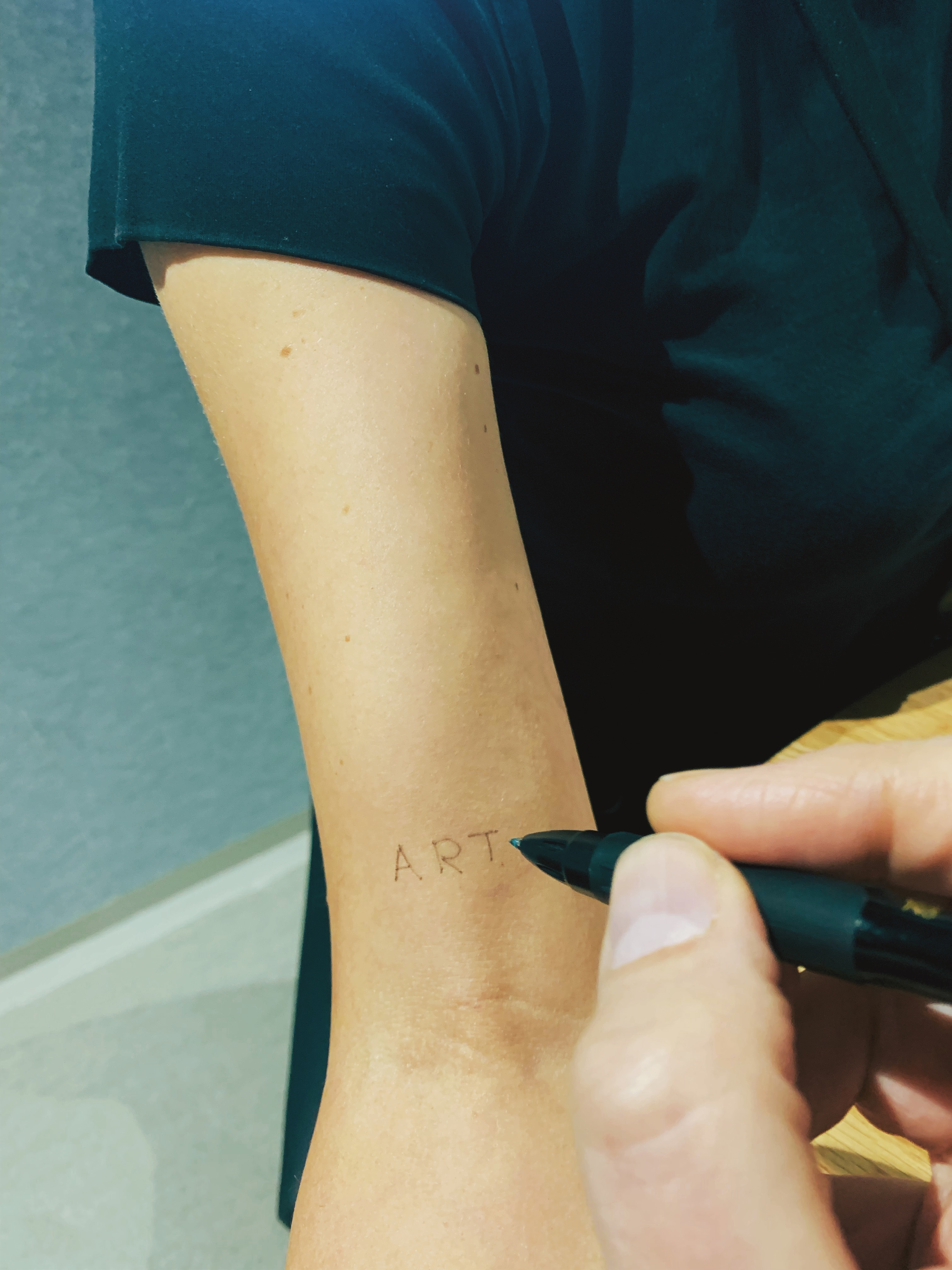 A hand holds a pen over someone else's bicep, on which the word ART has been written