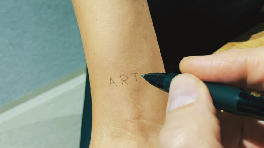 A hand holds a pen over someone else's bicep, on which the word ART has been written