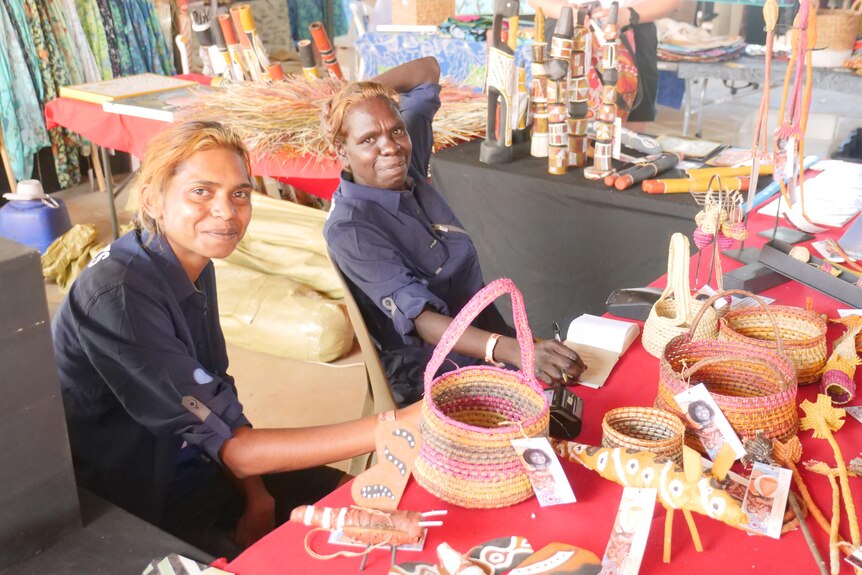 Two women sitting at a table covered in woven baskets and other traditional Indigenous artworks.
