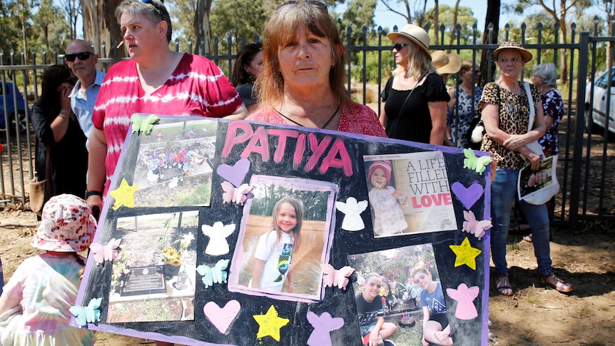 A woman at a cemetery holding a large poster bearing the name "Patiya", which is covered in photos and news clippings.