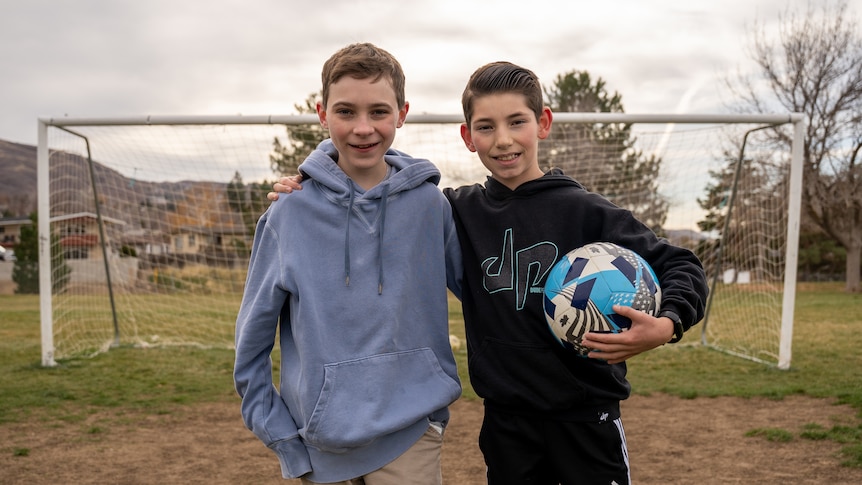 Two young boys stand close together in a soccer field, smiling