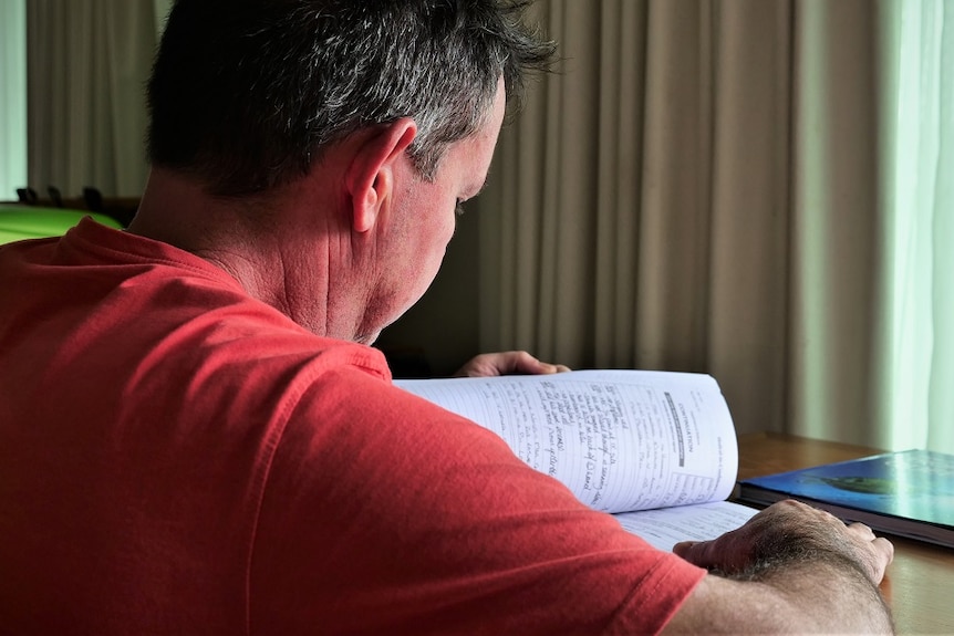 Middle-aged man in red shirt looking through medical documents at a small table in a hotel room.
